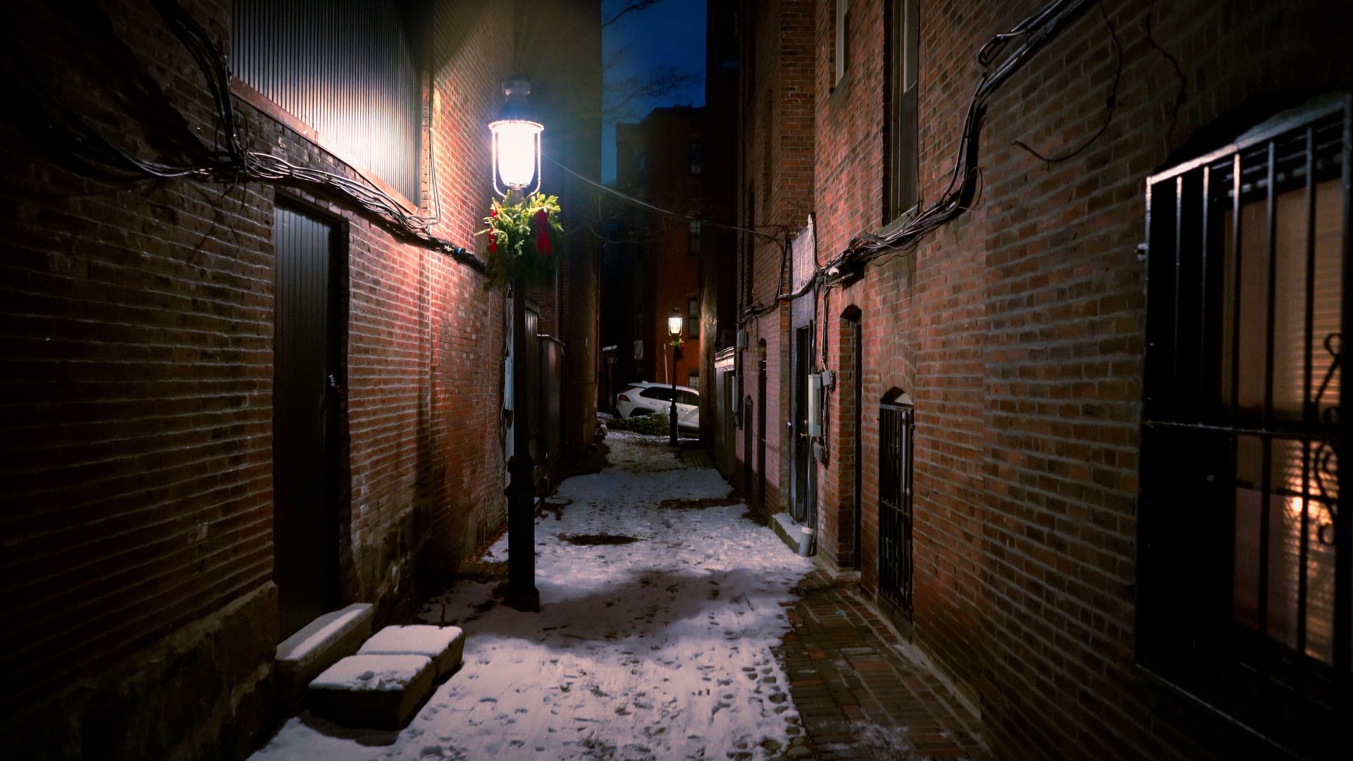 Beacon Hill at midnight in winter snow: a lantern hung with holiday greenery above a footprinted snowy alleyway.