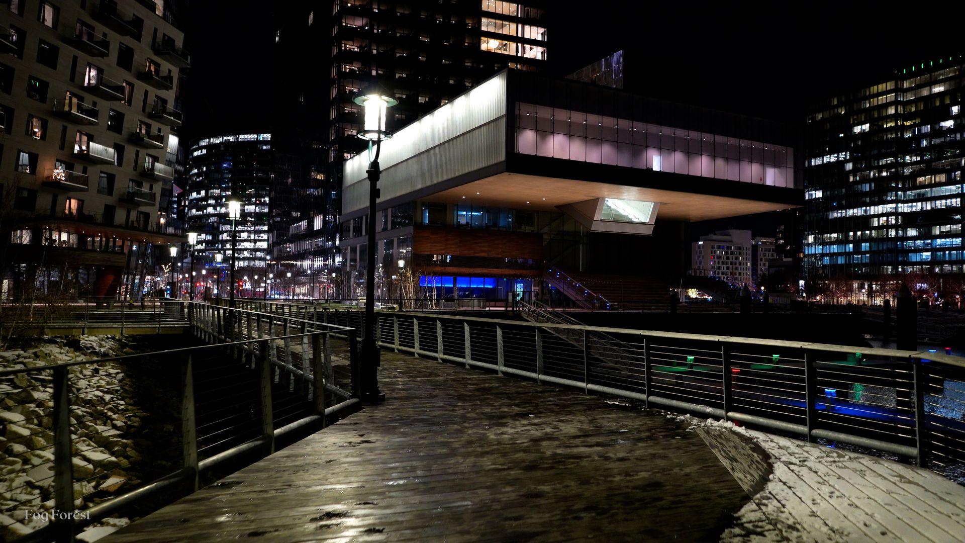 Boston Seaport at midnight in freezing cold: patches of snow on a boardwalk beneath a lit cantilevered building
