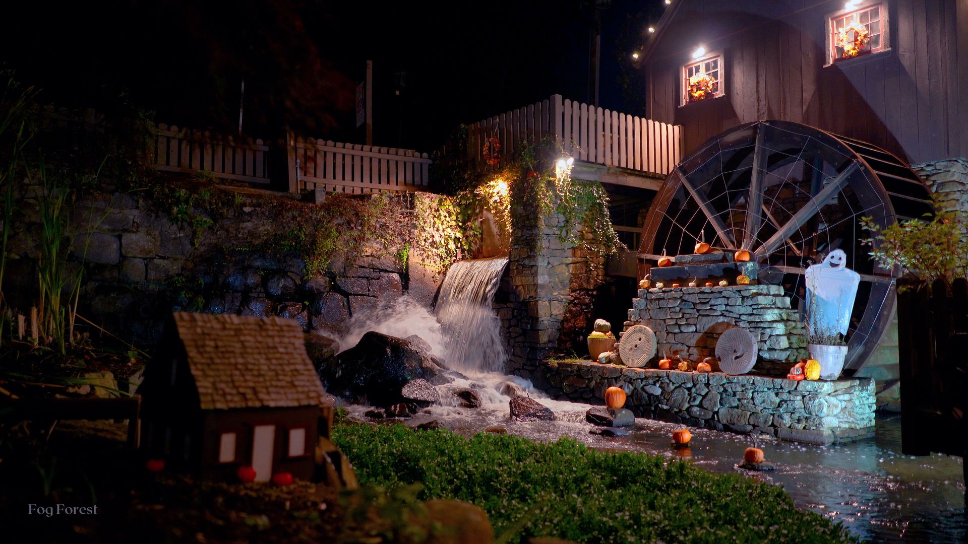 Plymouth Grist Mill at night on Halloween: a floodlit waterfall churning beside the wooden waterwheel, pumpkins set along wet stone steps.