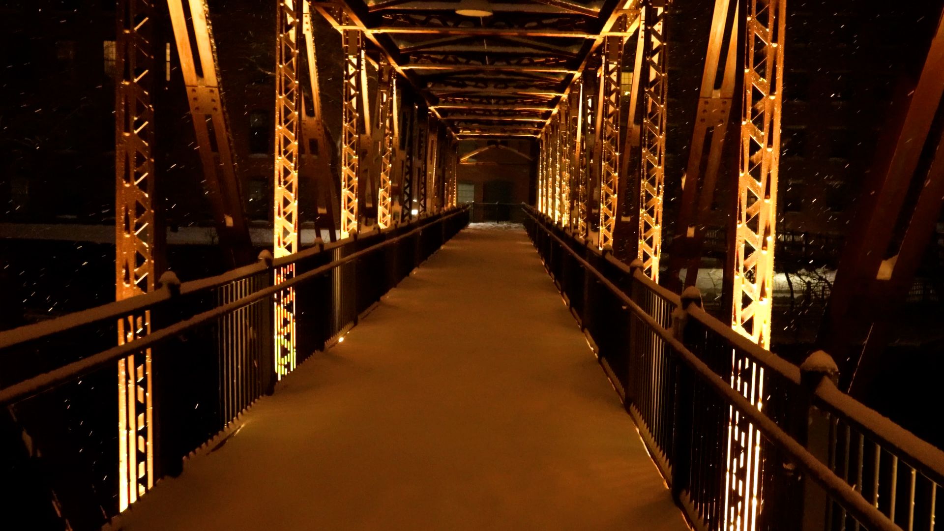 Snow falling through the lit trusses of an iron footbridge at night