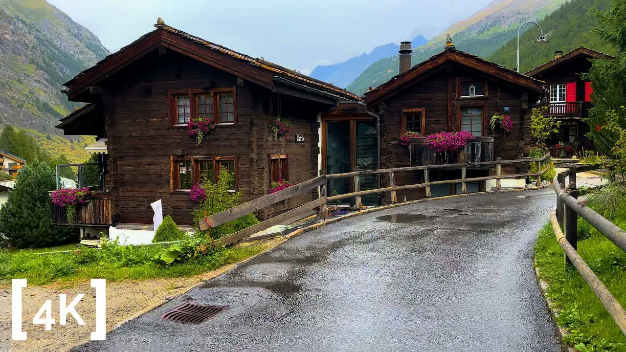 Zermatt on a rainy day: dark timber chalets with pink flower boxes above a wet lane, cloud low on the mountains.
