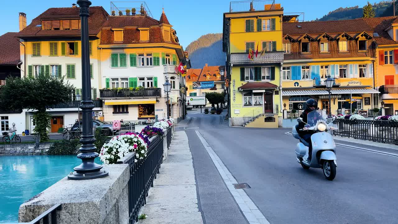 Interlaken at sunrise in clear early light: a stone bridge with flower boxes above emerald river water, shuttered facades catching the sun.