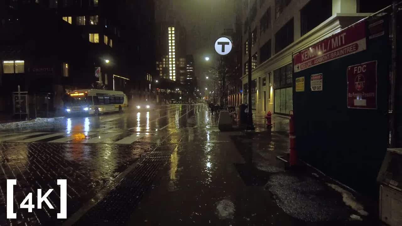 Kendall Square at midnight in rain: a lit red subway station sign above a wet, reflective sidewalk