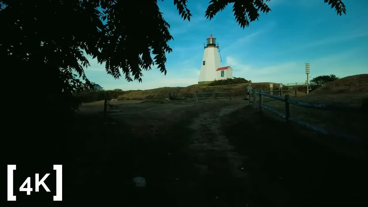 A New England lighthouse at daytime in clear weather: a white tower rising behind a split-rail fence at the end of a dirt path