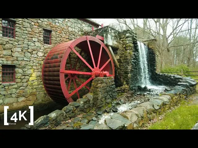 A New England grist mill in the evening after rain: a red water wheel on a fieldstone wall beside a narrow falls.