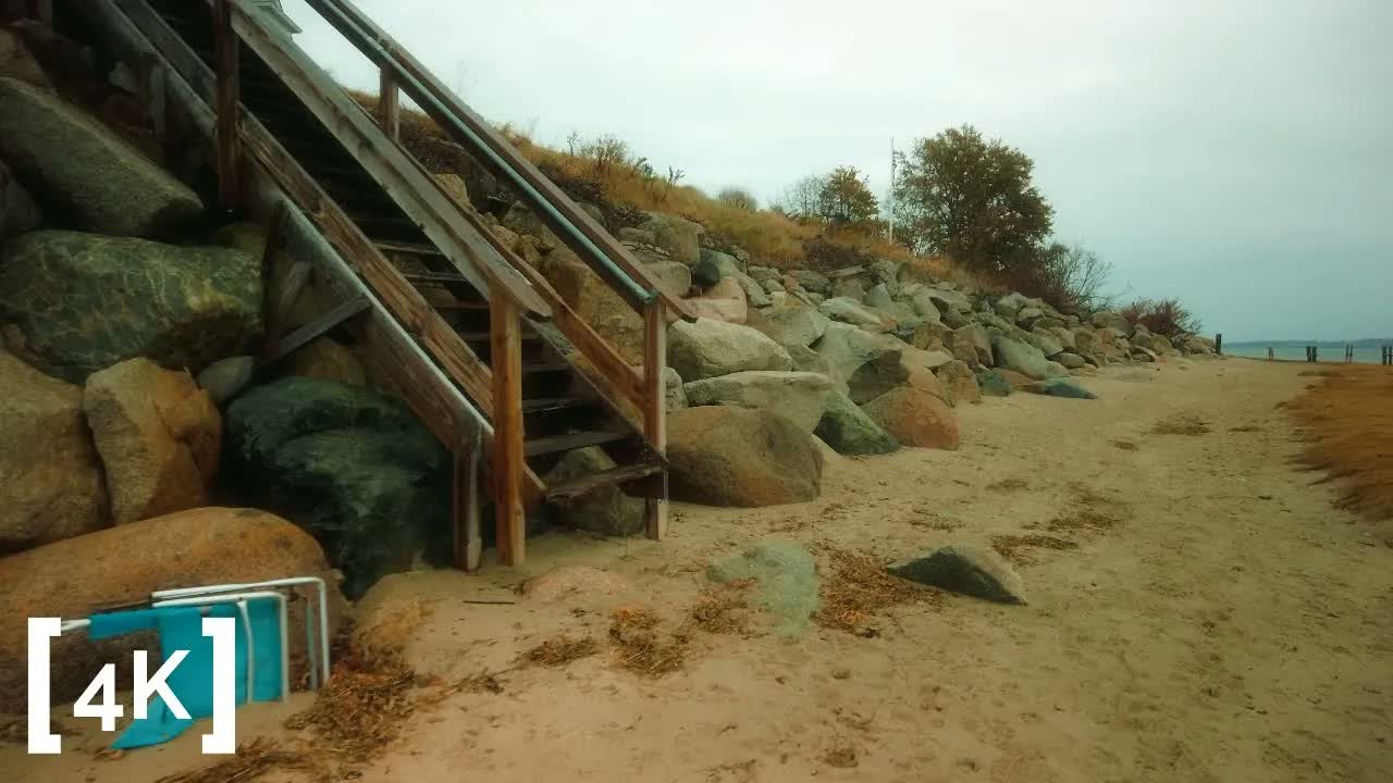 An East Coast beach at morning in rain: a wooden staircase over granite boulders, a folded beach chair tipped at its foot.
