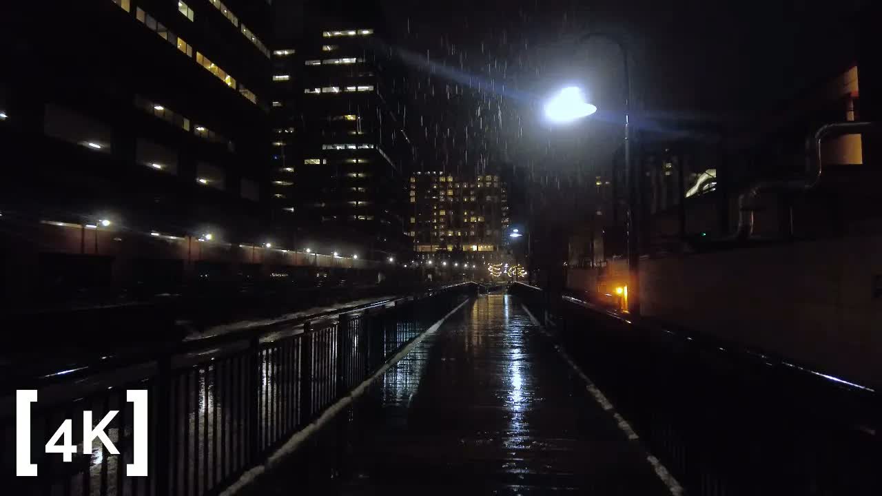 Cambridge Harborwalk at midnight in heavy rain: rain streaking past a streetlamp over a wet railed walkway.