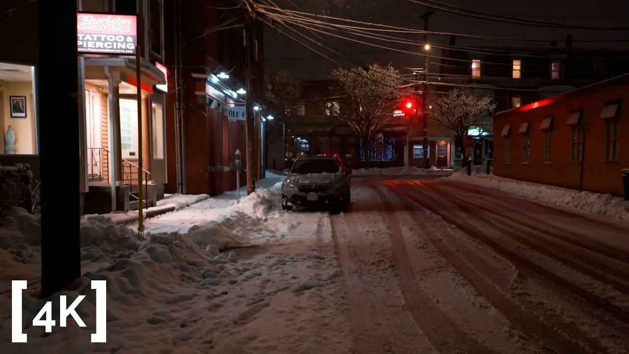 A town at night in a snowstorm: a parked car buried in fresh snow beside a lit shop sign