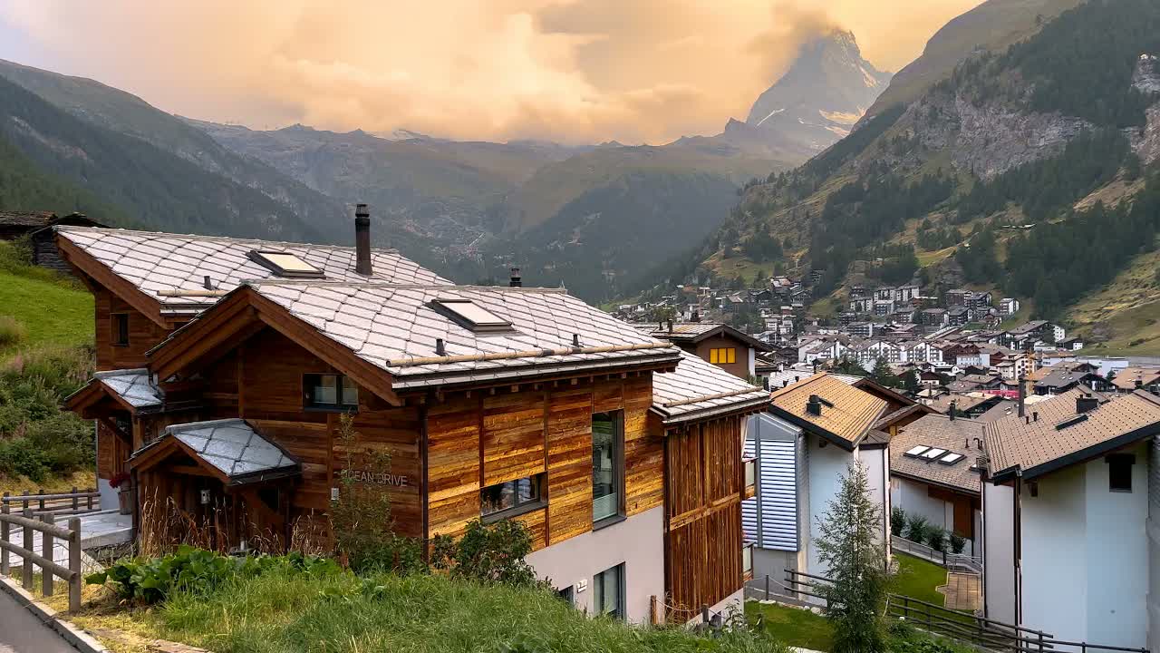 Zermatt, Switzerland at sunset in summer: stone-shingled chalet roofs above the village, the Matterhorn half-veiled in orange cloud.
