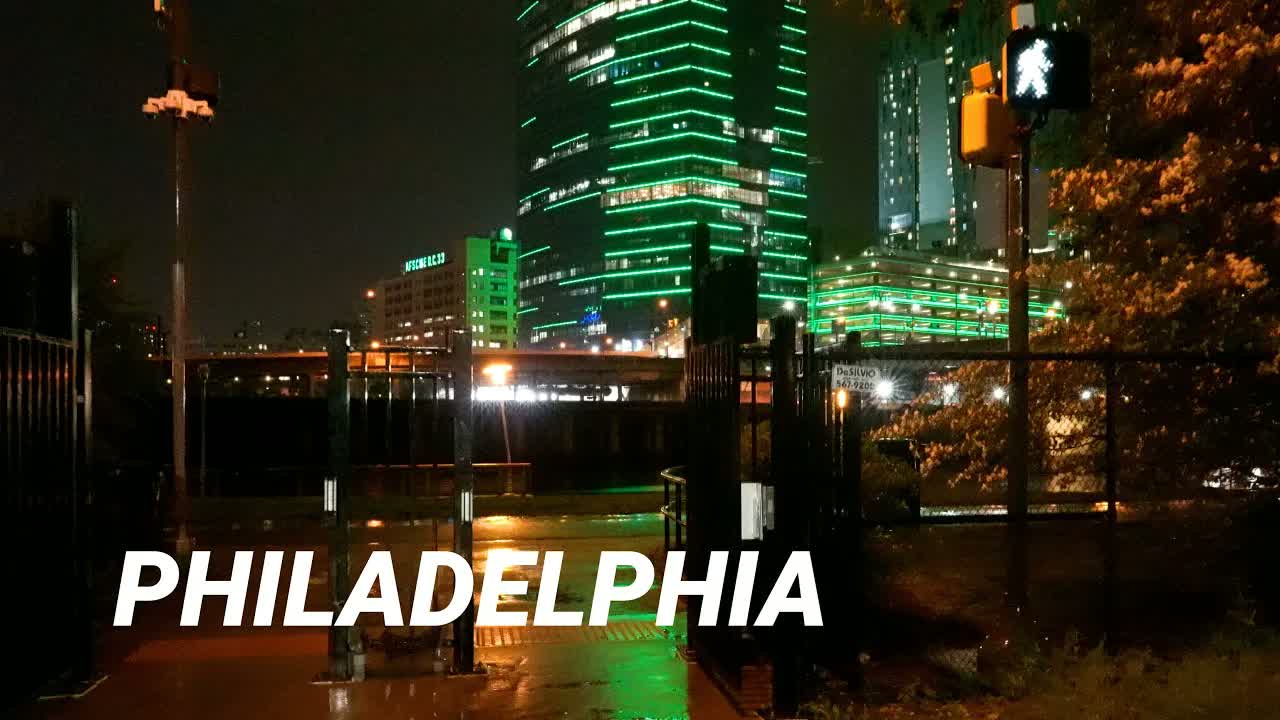 Philadelphia at night in rain: a tower banded in green light above a wet riverside path, a walk signal glowing beside the trees.