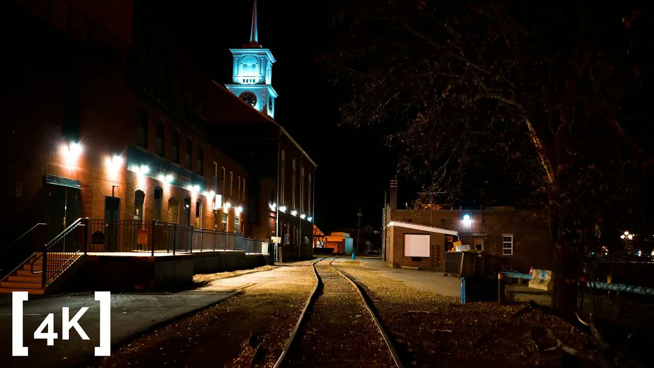 An old New England factory town at late night in lamplight: a blue-lit clock tower above brick mills and curving rail tracks.
