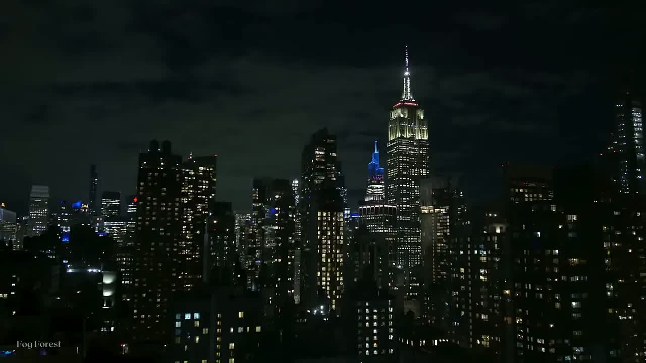 Manhattan at night from an open window: the Empire State Building lit white over a skyline of scattered lit windows.