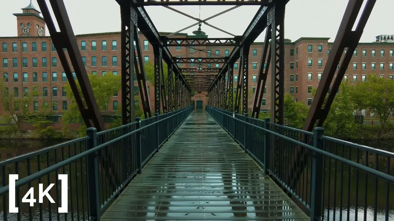 A New England mill town at afternoon after rain: a wet steel truss footbridge leading toward brick mill buildings with a clock tower