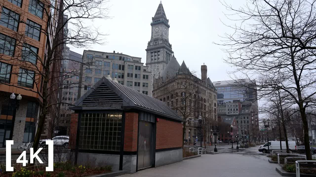 Boston in daytime rain: a stone clock tower rising above bare trees and a wet, nearly empty brick plaza.