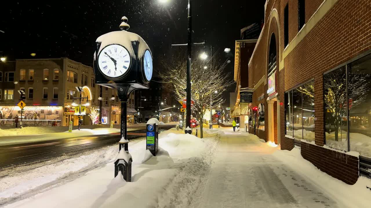 A New England town at night in snow: a black street clock capped with snow beside a snowbanked sidewalk.