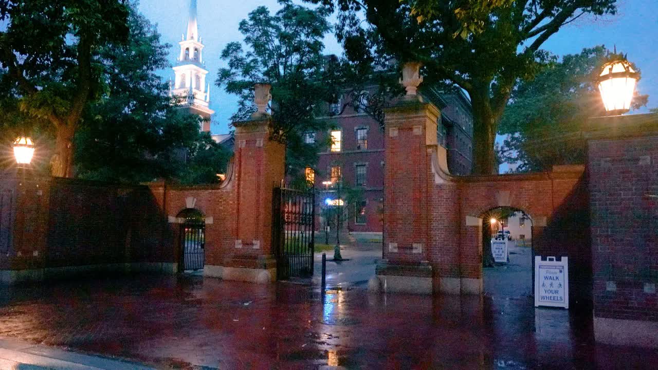 Harvard University, Cambridge at evening in rain: brick gate arches with lit lanterns, a white steeple rising behind wet trees.