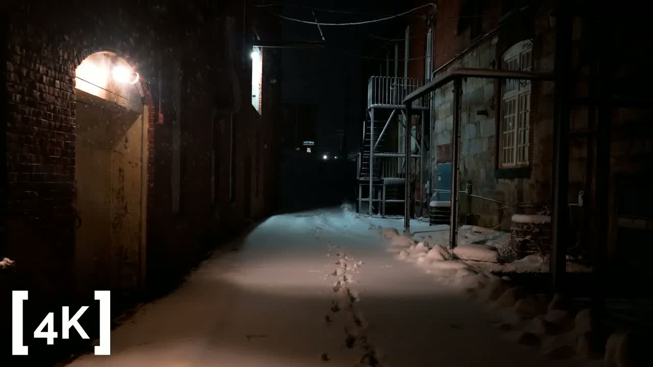 An old industrial park at late night in falling snow: a single line of footprints down an unplowed brick alley.