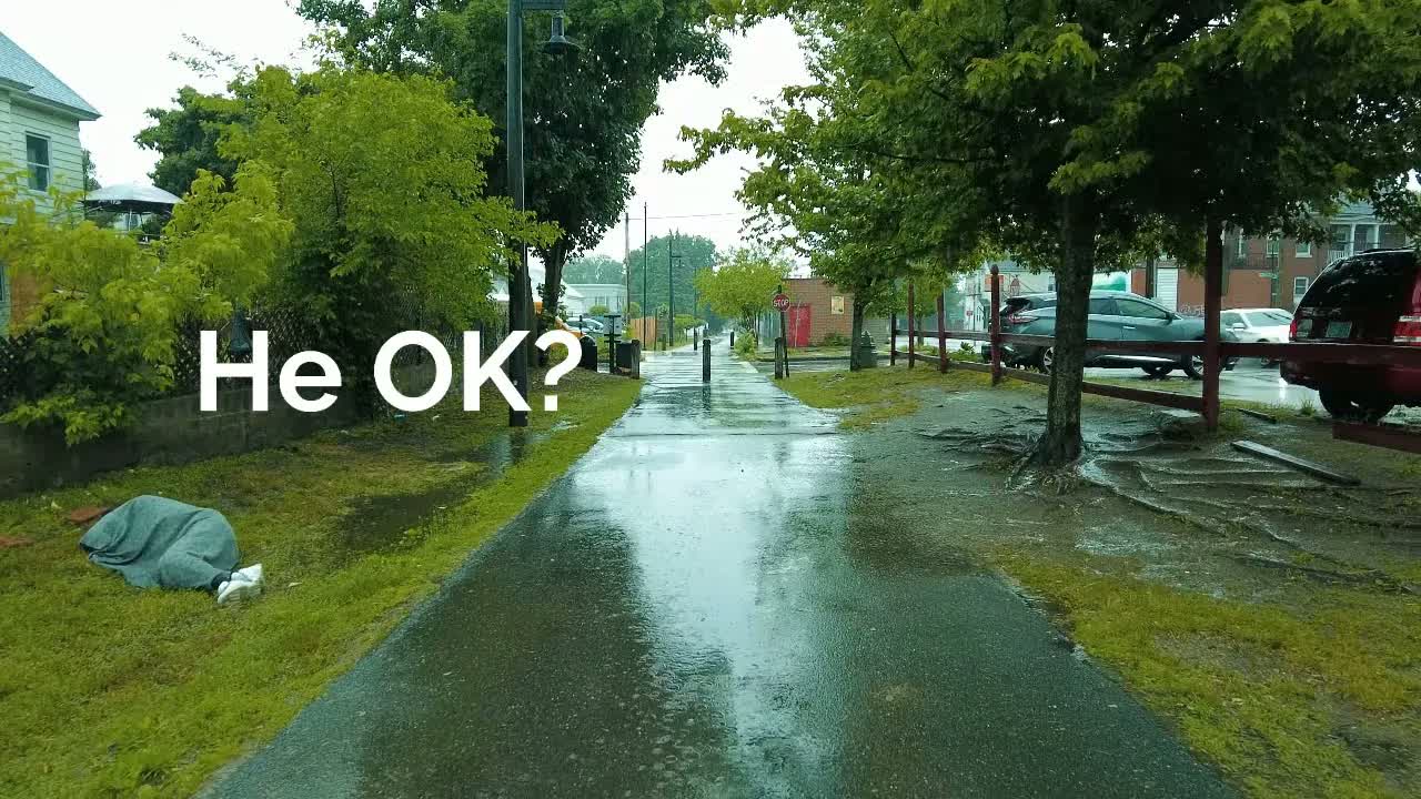 A New England mill town at early morning in heavy rain: a wet rail-trail path running between soaked trees and parked cars.
