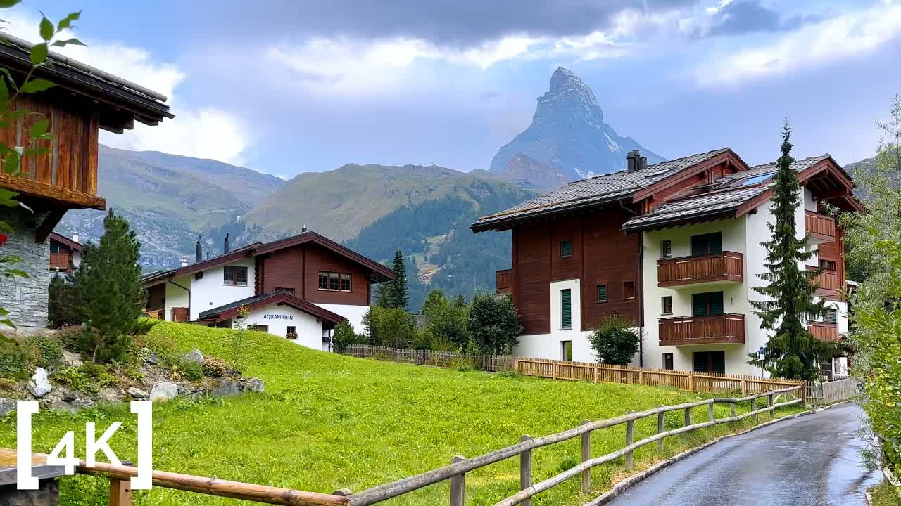 Zermatt at daytime in rain: the Matterhorn above chalet balconies and a wet lane along a wooden rail fence
