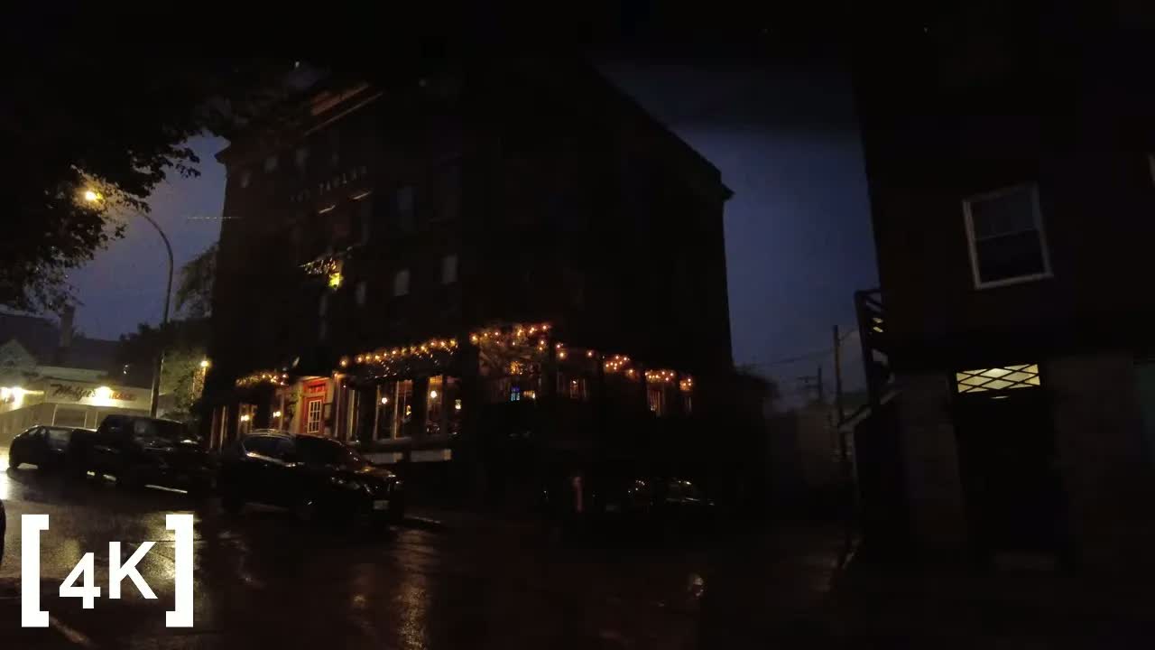 A restaurant corner at night in heavy rain: strings of lights along dark windows above parked cars on a wet street.