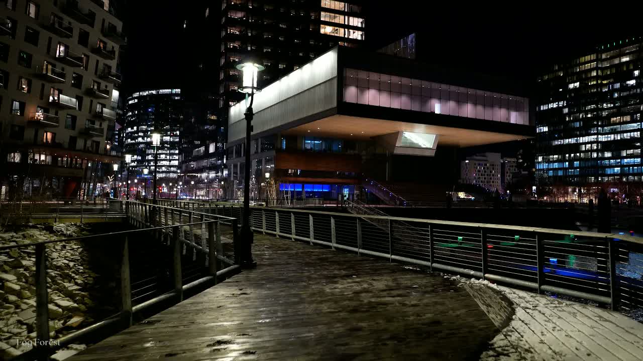 Boston Seaport at midnight in freezing cold: patches of snow on a boardwalk beneath a lit cantilevered building
