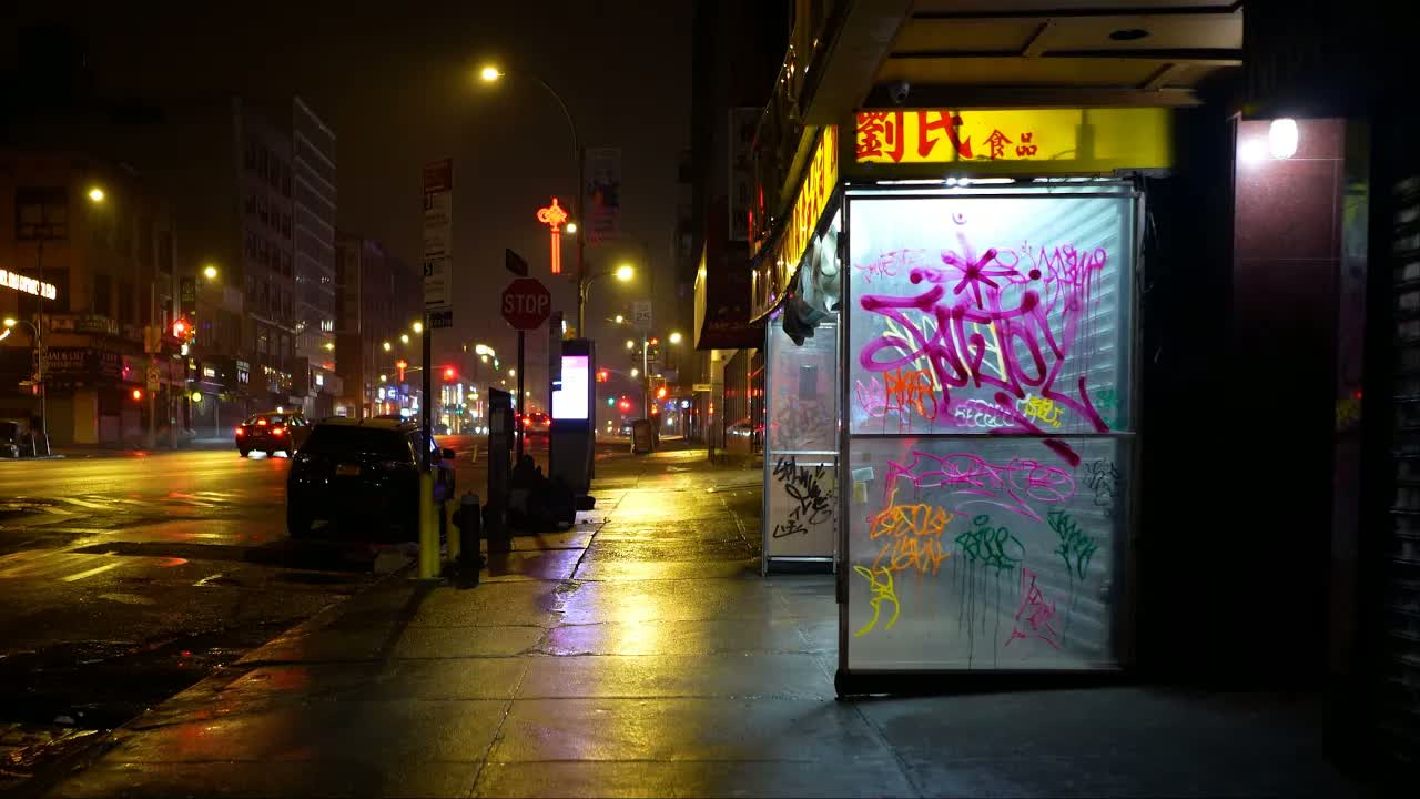 New York's Chinatown at 2 a.m. in rain: graffiti-tagged shutters glowing under a yellow storefront sign, a stop sign down the block.