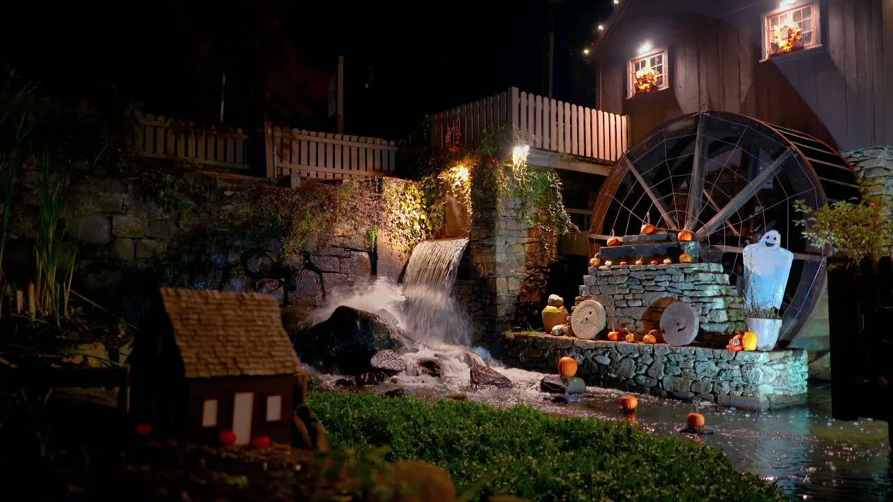 Plymouth Grist Mill at night on Halloween: a floodlit waterfall churning beside the wooden waterwheel, pumpkins set along wet stone steps.