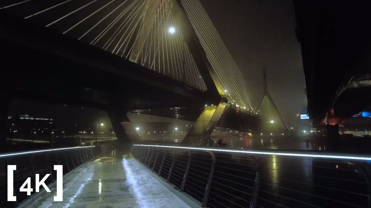 Zakim Bridge, Boston at 2 a.m. in rain: white cable stays lit against the dark, a glowing handrail strip along the wet footpath beneath.