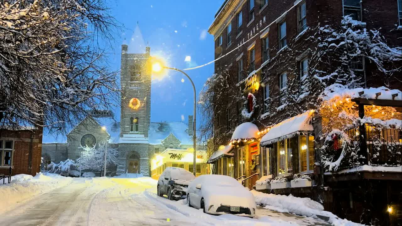A New England town at evening in fresh snow: a lit wreath on a stone church tower above snow-covered parked cars